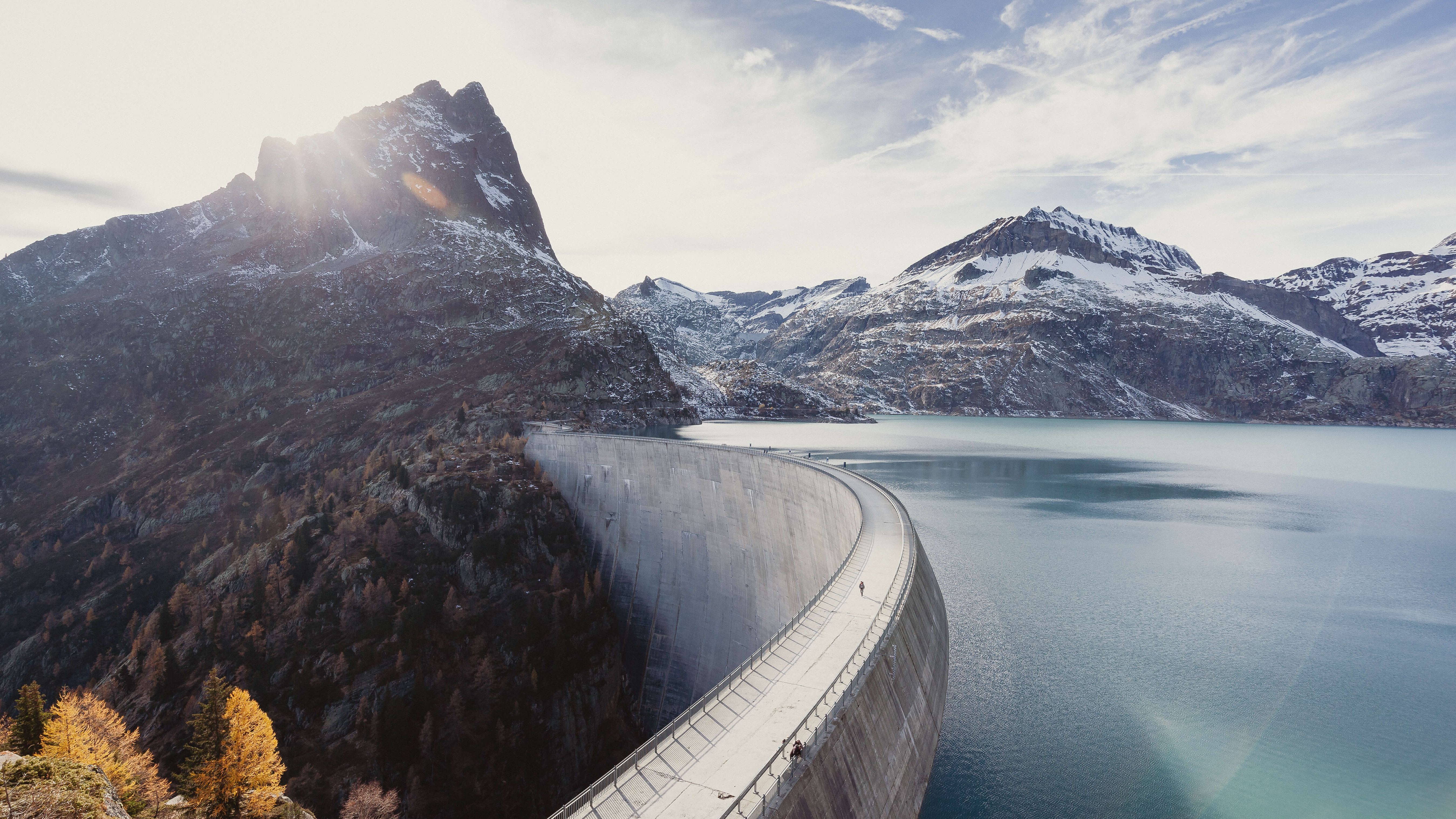 Eine grosse Staumauer in einem alpinen Tal, die sich halbkreisförmig durch die Landschaft zieht. Der türkisfarbene Stausee liegt eingebettet zwischen schneebedeckten Bergen. Im Hintergrund scheint die Sonne hinter einem markanten Berggipfel hervor.