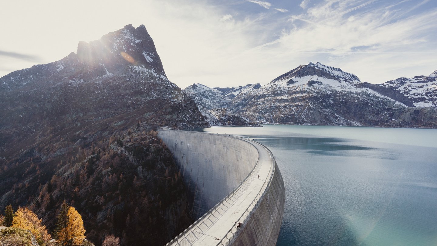Eine grosse Staumauer in einem alpinen Tal, die sich halbkreisförmig durch die Landschaft zieht. Der türkisfarbene Stausee liegt eingebettet zwischen schneebedeckten Bergen. Im Hintergrund scheint die Sonne hinter einem markanten Berggipfel hervor.
