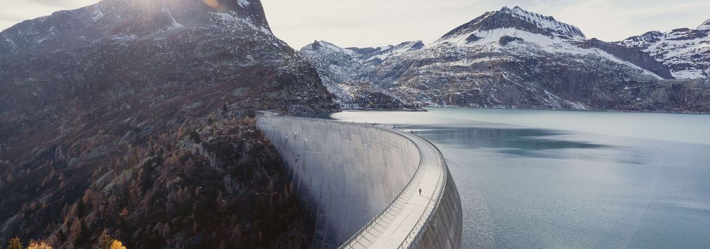 Eine grosse Staumauer in einem alpinen Tal, die sich halbkreisförmig durch die Landschaft zieht. Der türkisfarbene Stausee liegt eingebettet zwischen schneebedeckten Bergen. Im Hintergrund scheint die Sonne hinter einem markanten Berggipfel hervor.