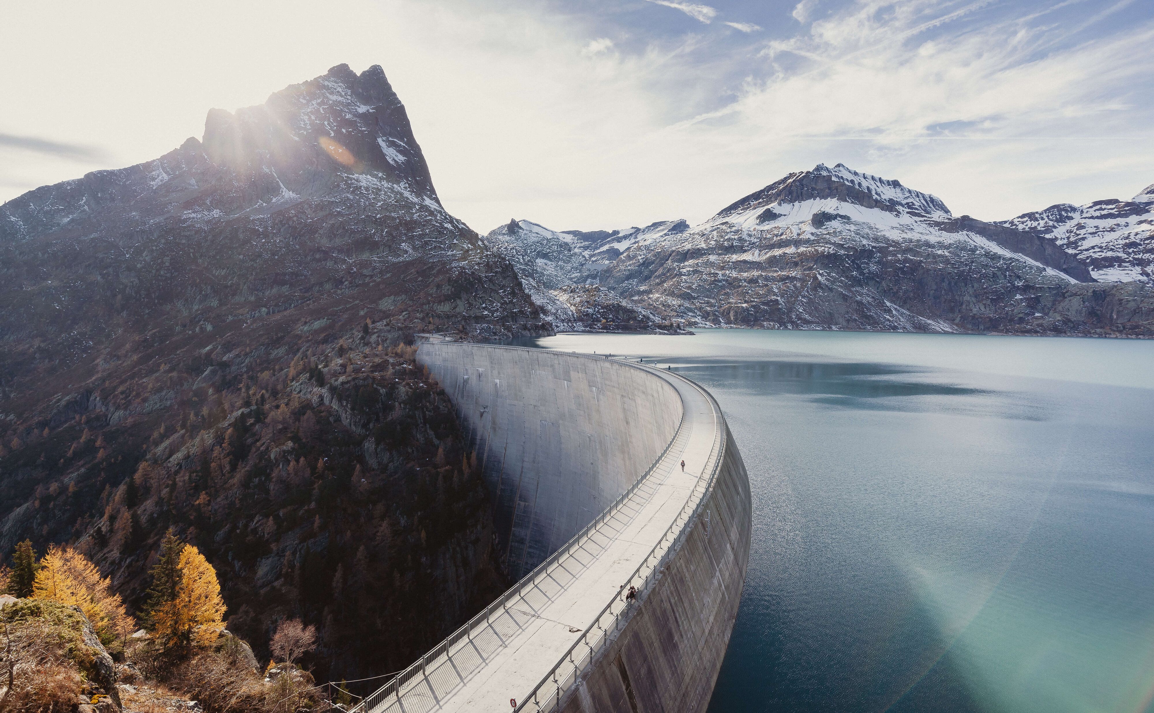 Eine grosse Staumauer in einem alpinen Tal, die sich halbkreisförmig durch die Landschaft zieht. Der türkisfarbene Stausee liegt eingebettet zwischen schneebedeckten Bergen. Im Hintergrund scheint die Sonne hinter einem markanten Berggipfel hervor.