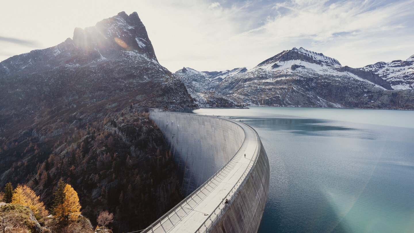 Eine grosse Staumauer in einem alpinen Tal, die sich halbkreisförmig durch die Landschaft zieht. Der türkisfarbene Stausee liegt eingebettet zwischen schneebedeckten Bergen. Im Hintergrund scheint die Sonne hinter einem markanten Berggipfel hervor.
