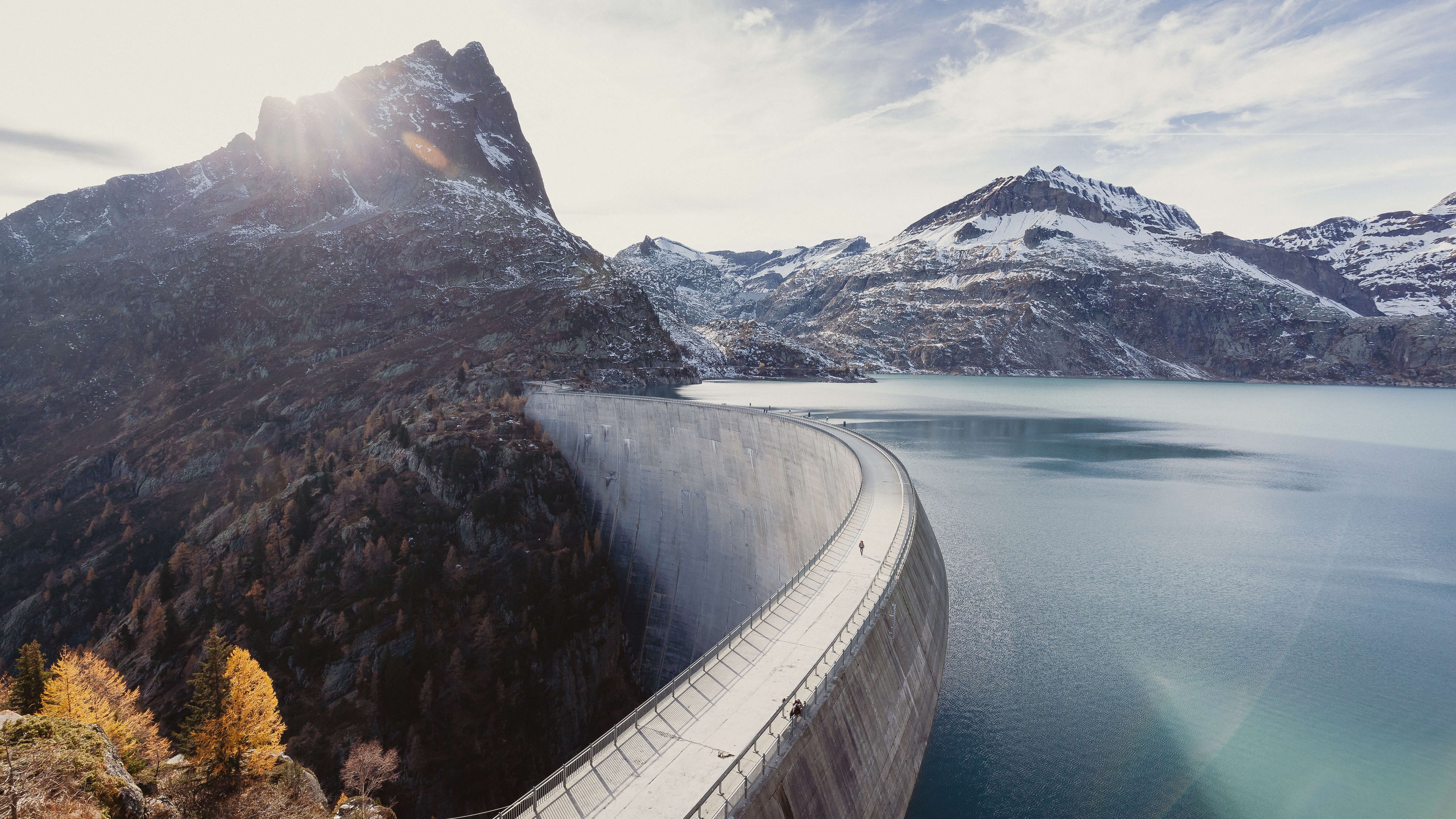 Eine grosse Staumauer in einem alpinen Tal, die sich halbkreisförmig durch die Landschaft zieht. Der türkisfarbene Stausee liegt eingebettet zwischen schneebedeckten Bergen. Im Hintergrund scheint die Sonne hinter einem markanten Berggipfel hervor.