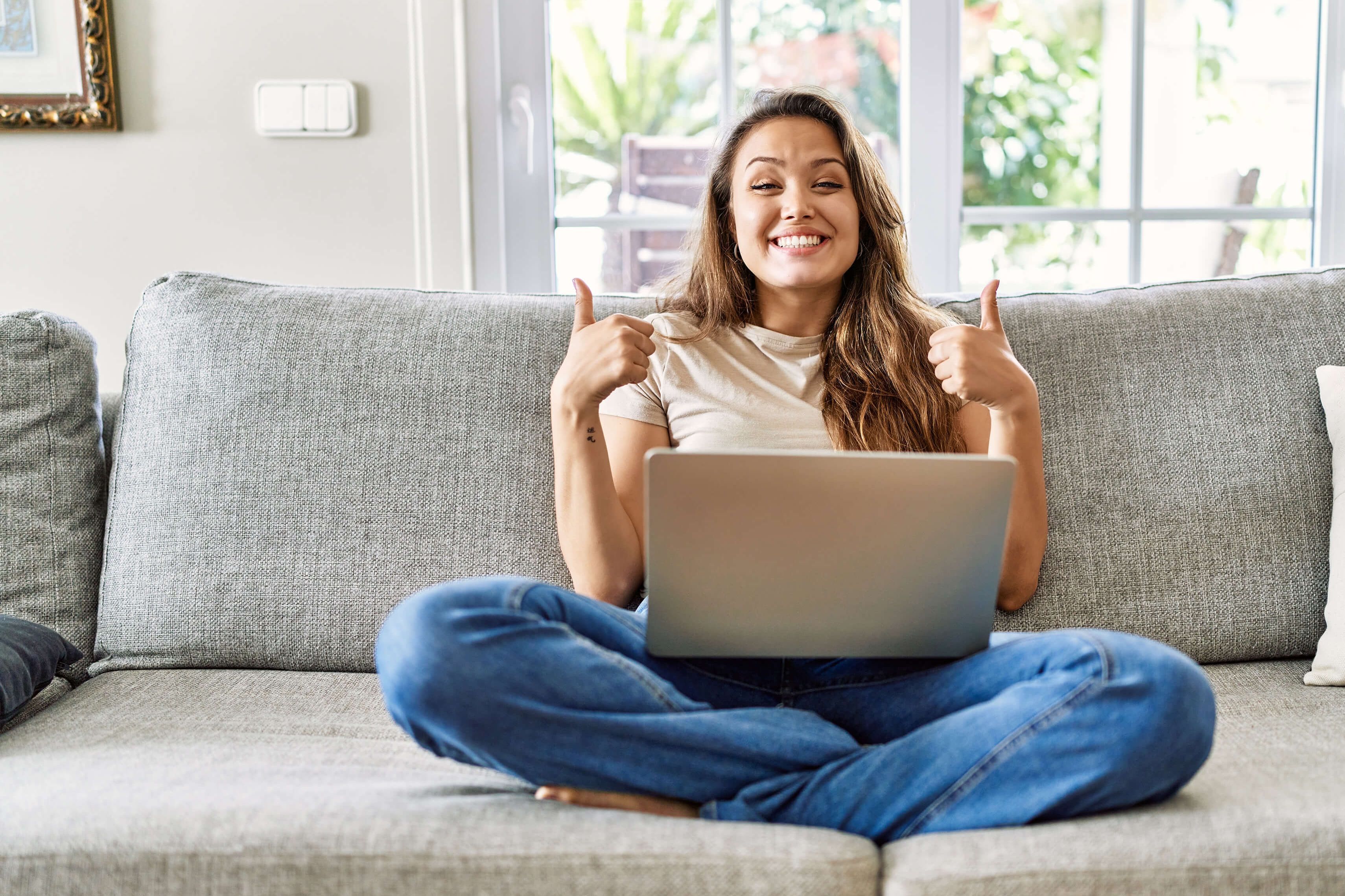 Une jeune femme est assise en tailleur sur le canapé avec son ordinateur portable sur les jambes et sourit à la caméra avec les deux pouces vers le haut