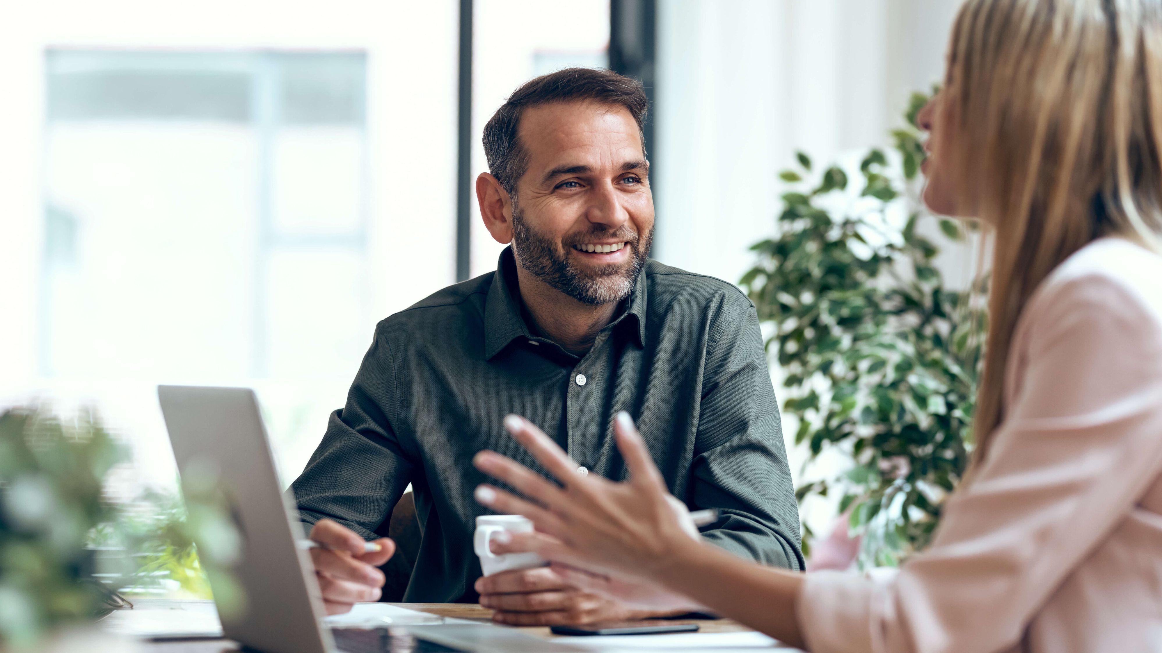 Man and woman sitting at a table in conversation, with the man visible from the front and the woman holding a laptop in front of her.