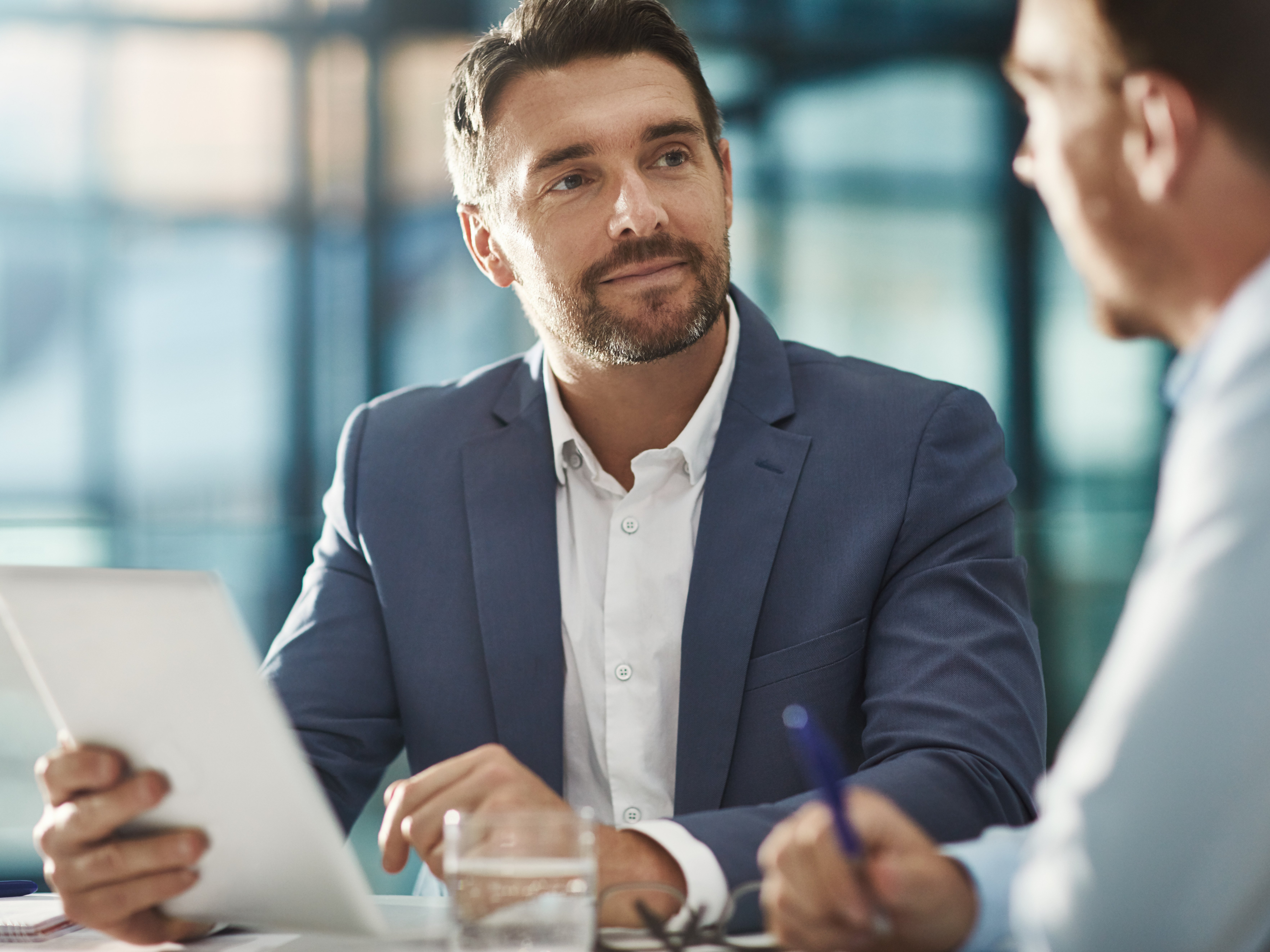Ein Mann im Anzug und weissem Hemd schaut im Rahmen eines Beratungsgespräches rüber zu einem anderen Mann und hört aufmerksam zu
