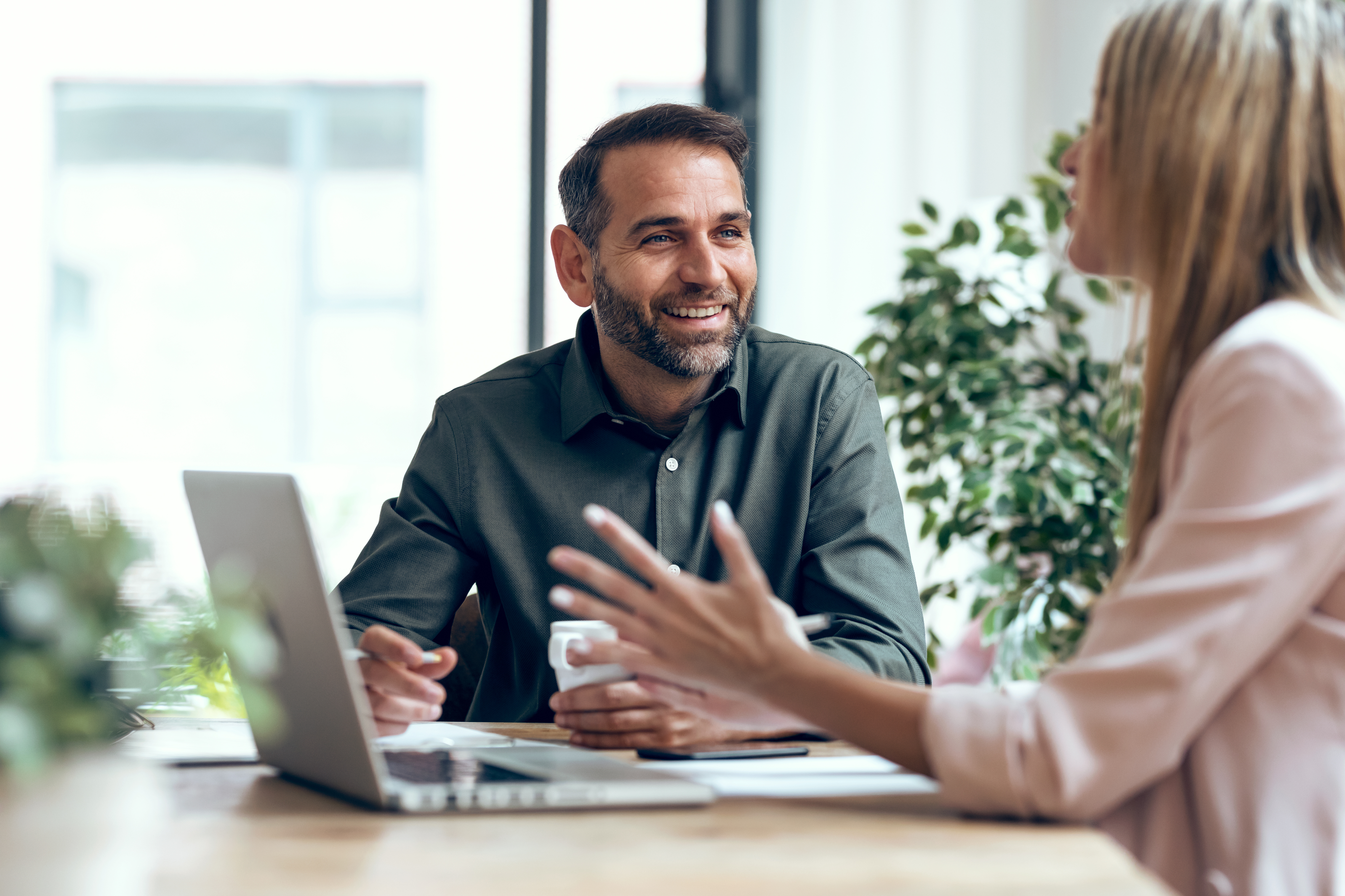 Man and woman sitting at a table in conversation, with the man visible from the front and the woman holding a laptop in front of her.