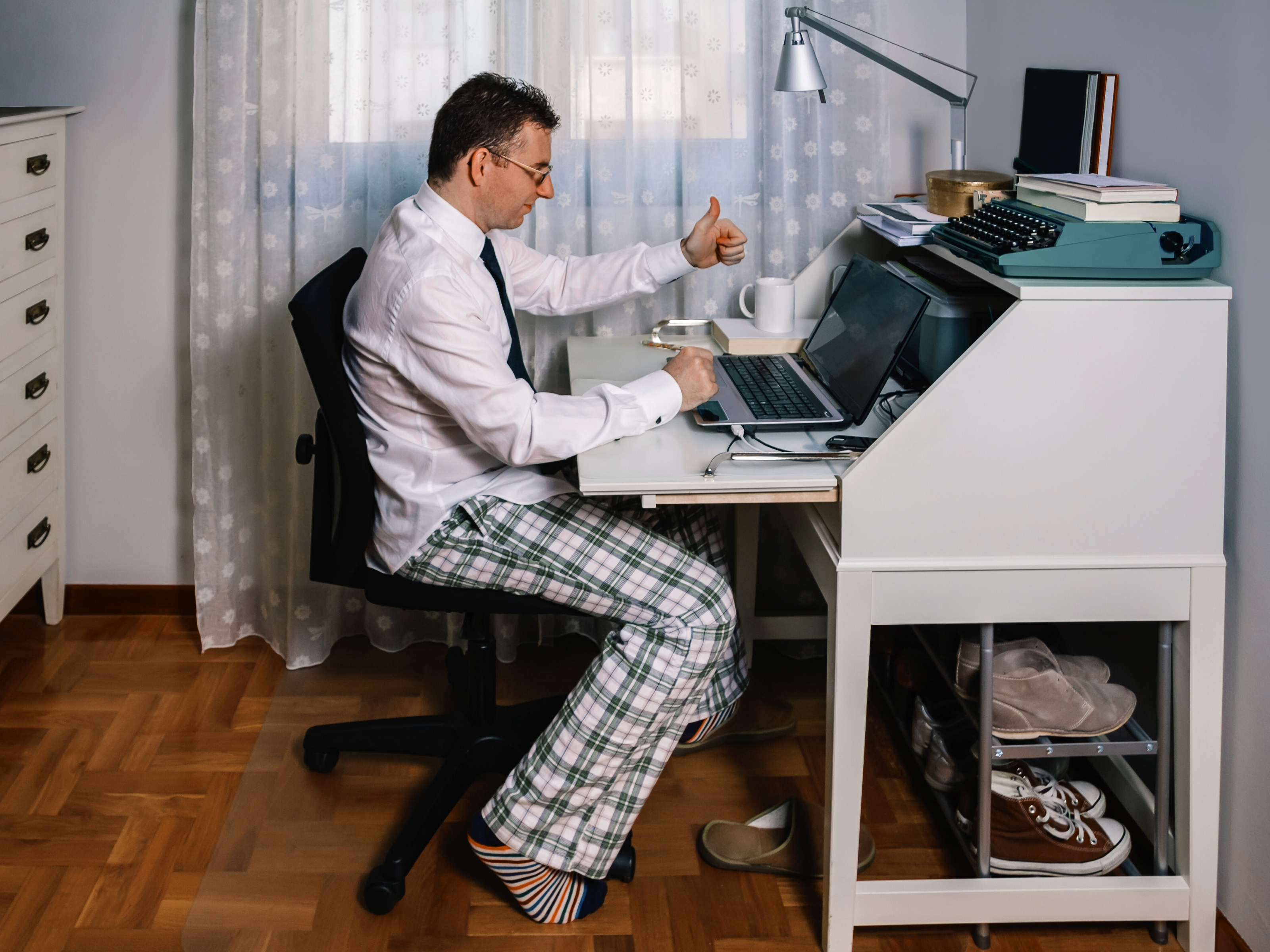 Mann im Homeoffice mit Hemd und Krawatte, aber Pyjamahose, sitzt am Schreibtisch und zeigt Daumen hoch in eine Laptop-Kamera.